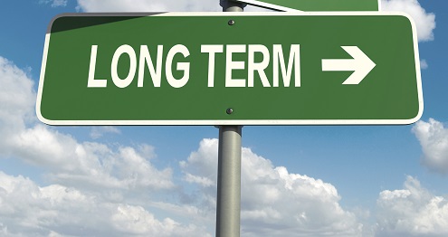 Road sign with "Long Term" written in white text on a green background, pointing to the right under a blue sky with clouds.